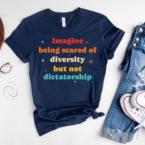 May include: Navy blue t-shirt with the text "Imagine being scared of diversity but not dictatorship" in colorful letters. The shirt is paired with blue jeans and white sneakers.