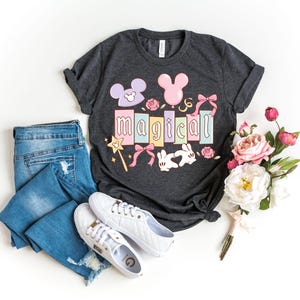 May include: Dark gray t-shirt with the word "MAGICAL" in pastel colors, featuring Mickey Mouse ears, bows, and a magic wand. The image also includes blue jeans, white sneakers, and a bouquet of flowers.