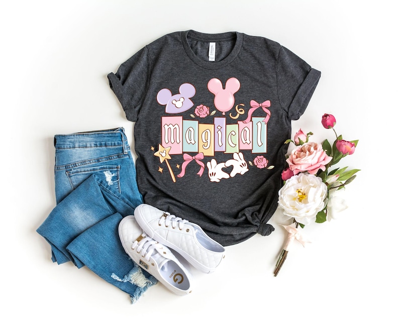 May include: Dark gray t-shirt with the word "MAGICAL" in pastel colors, featuring Mickey Mouse ears, bows, and a magic wand. The image also includes blue jeans, white sneakers, and a bouquet of flowers.
