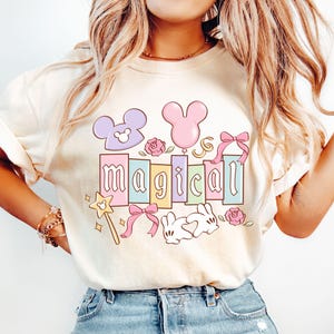 May include: Cream-colored t-shirt with a colorful "magical" graphic. The design includes Mickey Mouse-shaped balloons, roses, a magic wand, and a sleeping bunny. The word "magical" is spelled out in pastel-colored blocks.