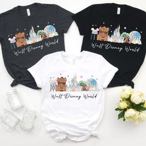 May include: Three t-shirts in charcoal, black, and white, each featuring a colorful graphic of a theme park skyline with the text "Walt Disney World". The design includes a castle, a tree, and a globe.