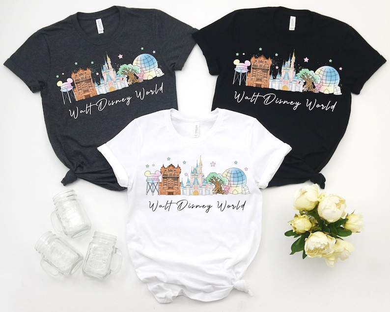 May include: Three t-shirts in charcoal, black, and white, each featuring a colorful graphic of a theme park skyline with the text "Walt Disney World". The design includes a castle, a tree, and a globe.