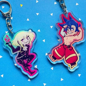 May include: Two colorful character keychains. One keychain features a character with blonde hair, wearing a black outfit and holding a flaming object. The other keychain features a character with blue hair, wearing red pants and a white shirt. Both keychains are made of clear acrylic.