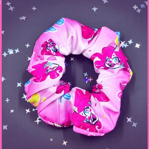 May include: Pink and white scrunchie with a repeating pattern of a cartoon character with pink hair, a party hat, and a big smile. The character is holding a balloon.