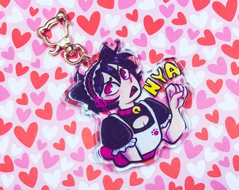 Op de afbeelding: Een zwarte en roze anime-sleutelhanger met een personage met zwart haar, rode ogen en een dienstmeidenoutfit. Het personage houdt een hand omhoog met het woord "NYA" erop geschreven. De sleutelhanger heeft een gouden metalen ring.