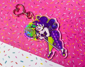 Peut inclure: Un charme holographique représentant un personnage de dessin animé avec des cheveux violets, un costume vert et violet et une sucette. Le personnage tient une baguette avec une étoile à l'extrémité. Le charme est attaché à un porte-clés argenté.
