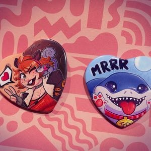 May include: Two heart-shaped buttons with cartoon illustrations. One features a red-haired character with a rat, the other a blue shark with the word "MRRR". Both are on a pink background with a repeating pattern.