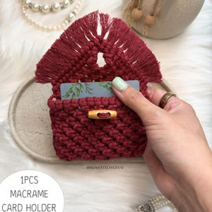 May include: A dark red macrame card holder featuring a triangular flap with fringe, a knotted body, and a horizontal wooden toggle closure. A grey card with a green leaf pattern is inserted inside. The text 1PCS MACRAME CARD HOLDER is visible.