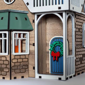 May include: A wooden model of a house with a green roof, a blue door with a green wreath, and brick details. The house has a porch with white railings and a window with a red bow.