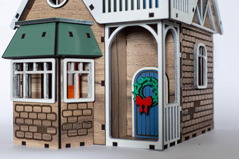 May include: A wooden model of a house with a green roof, a blue door with a green wreath, and brick details. The house has a porch with white railings and a window with a red bow.