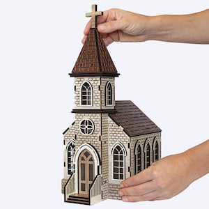 K&ouml;nnte beinhalten: Ein Holzmodell einer Kirche mit einem braunen Dach und einem Kreuz auf der Spitze. Die Kirche hat eine Steinfassade mit Fenstern und einer T&uuml;r. Das Modell wird von einer Hand gehalten.