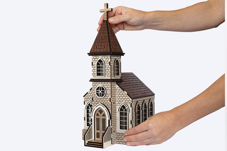 K&ouml;nnte beinhalten: Ein Holzmodell einer Kirche mit einem braunen Dach und einem Kreuz auf der Spitze. Die Kirche hat eine Steinfassade mit Fenstern und einer T&uuml;r. Das Modell wird von einer Hand gehalten.