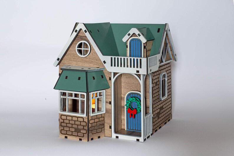 May include: A wooden model house with a green roof, white trim, and a blue door. The house has a small balcony with a white railing and a wreath on the door.