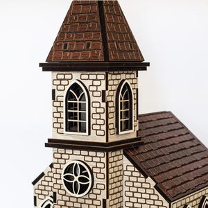 K&ouml;nnte beinhalten: Ein Holzmodell einer Kirche mit einem braunen Dach und wei&szlig;en Backsteinmauern. Die Kirche hat einen hohen Turm mit einem spitzen Dach und zwei Bogenfenstern. Im unteren Bereich des Turms befindet sich ein rundes Fenster mit einem dekorativen Muster.