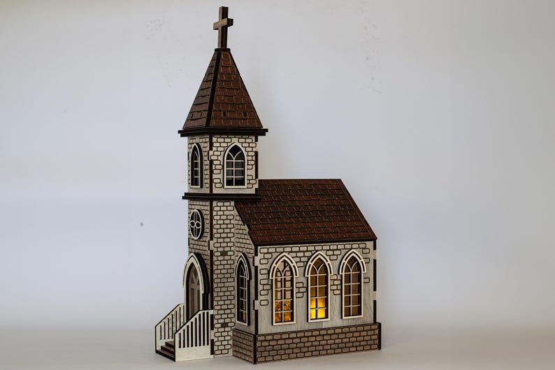 K&ouml;nnte beinhalten: Ein Holzmodell einer Kirche mit braunem Dach, wei&szlig;en Backsteinw&auml;nden und Fenstern, durch die Licht scheint. Die Kirche hat einen hohen Turm mit einem Kreuz auf der Spitze.
