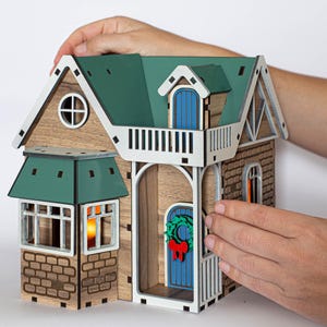 May include: A wooden model of a house with a green roof, a blue door, and a wreath on the door. The house has a balcony and a brick facade. The model is being held by a person's hands.