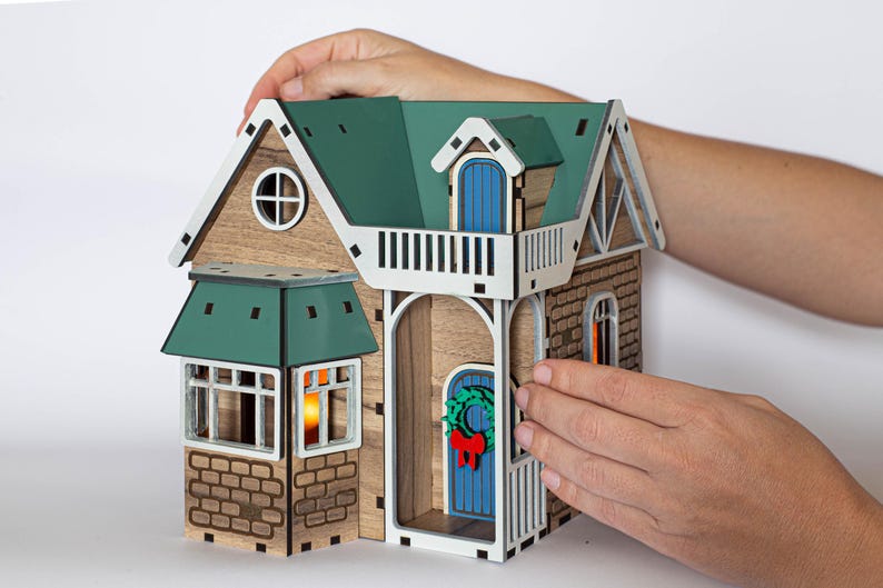 May include: A wooden model of a house with a green roof, a blue door, and a wreath on the door. The house has a balcony and a brick facade. The model is being held by a person's hands.