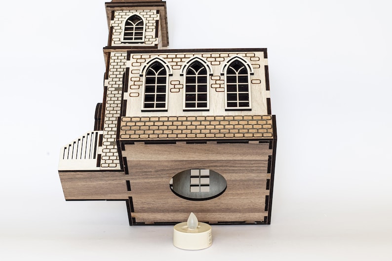 K&ouml;nnte beinhalten: Ein Holzmodell einer Kirche mit brauner gebeizter Oberfl&auml;che. Die Kirche hat drei Fenster und eine kleine Veranda. Ein wei&szlig;es LED-Teelicht steht vor der Kirche.