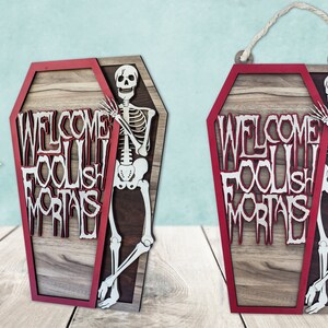 May include: Wooden coffin-shaped sign with a red border and the words "WELCOME FOOLISH MORTALS" in white. A skeleton figure is positioned on the right side. The sign includes a rope for hanging.