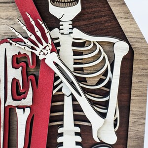 May include: A layered wooden Halloween decoration featuring a skeleton and the word "WELCOME". The skeleton is light-coloured, with a red accent and a dark brown background.