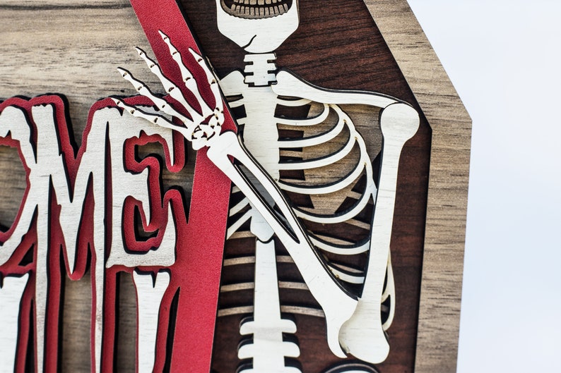 May include: A layered wooden Halloween decoration featuring a skeleton and the word "WELCOME". The skeleton is light-coloured, with a red accent and a dark brown background.