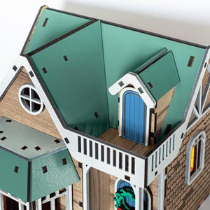May include: A wooden model of a house with a green roof, white trim, and a blue door. The house has a small balcony and a window with a wreath.