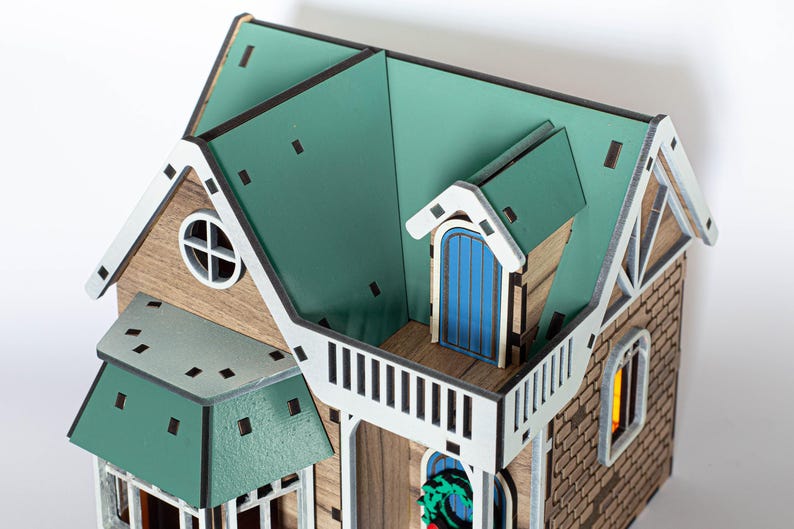 May include: A wooden model of a house with a green roof, white trim, and a blue door. The house has a small balcony and a window with a wreath.