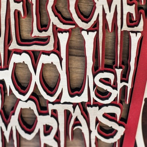 May include: A layered wooden sign with the words "WELCOME FOOLISH MORTALS" cut out in a gothic font. The sign features red, black, and natural wood tones, with a skeleton design on the right side.