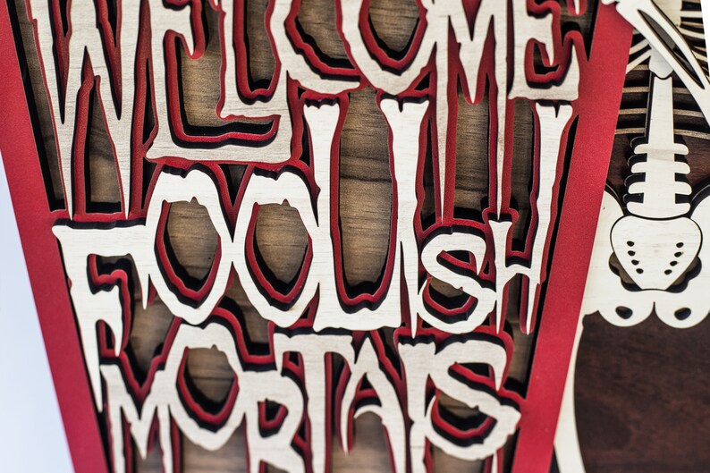 May include: A layered wooden sign with the words "WELCOME FOOLISH MORTALS" cut out in a gothic font. The sign features red, black, and natural wood tones, with a skeleton design on the right side.