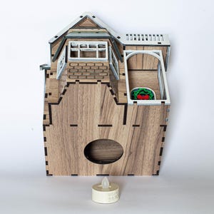May include: A wooden house-shaped candle holder with a white roof and a small window. The candle holder has a cutout in the front for a tea light candle. The candle holder is sitting on a white surface with a tea light candle in the cutout.