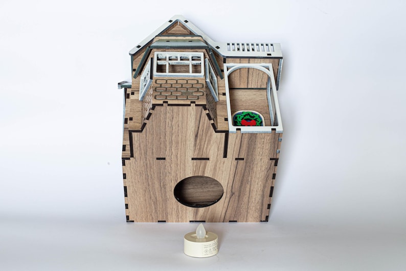May include: A wooden house-shaped candle holder with a white roof and a small window. The candle holder has a cutout in the front for a tea light candle. The candle holder is sitting on a white surface with a tea light candle in the cutout.