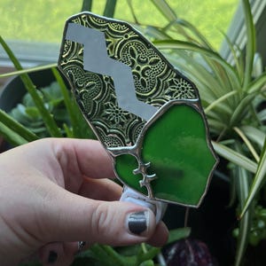 May include: A stained glass night light with a green and gold design. The night light has a silver border and a white plug. The green glass is shaped like a heart. The gold glass has a decorative pattern.