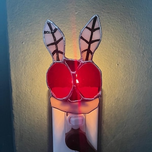 May include: A stained glass nightlight shaped like a bunny with red, white, and black accents. The nightlight is plugged into an electrical outlet.