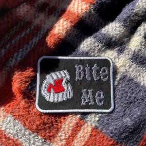 May include: Black and white embroidered patch with a red tooth and the text "Bite Me" in white.