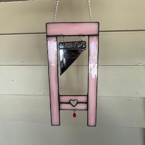 May include: Pink stained glass guillotine with a silver chain for hanging. The design includes a heart and a red teardrop. The blade is mirrored, reflecting the surroundings. The frame is a light pink color.