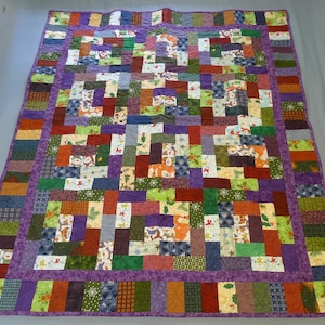 May include: A patchwork quilt with a vibrant mix of colors and patterns. The quilt features rectangular blocks in shades of purple, green, orange, and red, with a purple border. Some blocks have holiday-themed prints. The quilt is laid out on a neutral surface.