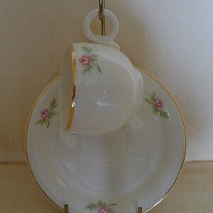 Peut inclure: Une tasse à thé et une soucoupe en porcelaine blanche avec une bordure dorée et un motif floral délicat de roses roses.