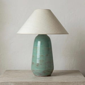 Op de afbeelding: Een turquoise keramische tafellamp met een crèmekleurige lampenkap. De lamp heeft een afgeronde voet en een gladde, glanzende afwerking. De lampenkap is conisch en gemaakt van een lichtgekleurde stof. De lamp staat op een steenkleurig oppervlak.