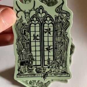 May include: A pale green sticker featuring a black ink illustration of a gothic arched window flanked by bookshelves filled with jars, plants, and a skull. A cat sits on the ledge near a mortar and pestle, with birds flying outside the window.