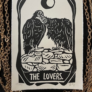 May include: Black and white linocut print of two vultures perched on a stone wall, with a crescent moon in the background. The print is titled "THE LOVERS."