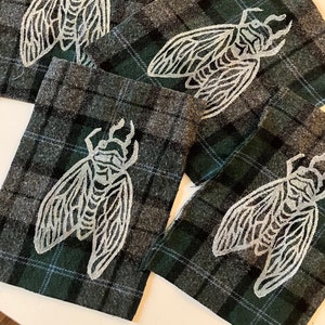 May include: Four fabric squares with a white cicada design on a green and gray plaid background.