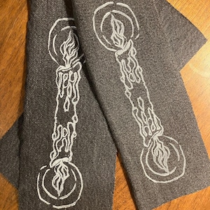 May include: Two gray fabric napkins with a white candle design. The candle illustration features a flame and dripping wax. The napkins are rectangular and appear to be made of a textured material.