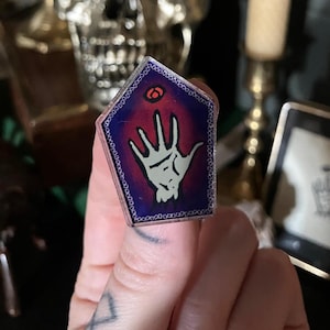 May include: A purple and red enamel pin with a white hand reaching up towards a red eye. The pin is shaped like a hexagon with a silver border.