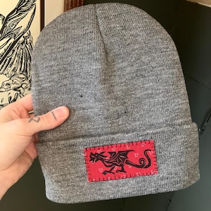 May include: A gray beanie with a red patch featuring a black dragon design. The patch is rectangular and sewn onto the folded brim of the hat. The beanie is made of a soft, knitted material.
