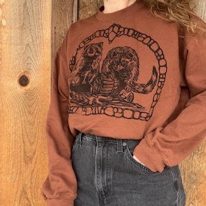 May include: A rust-colored sweatshirt featuring a black graphic of two raccoons inside a decorative border. The raccoons are depicted in a detailed, illustrative style. The sweatshirt is paired with dark wash denim jeans.