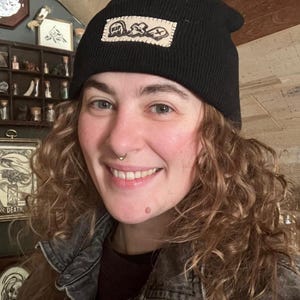 May include: Black beanie with a rectangular patch featuring an abstract design. The beanie is made of a knitted material. The patch has a white background with a stitched border. The beanie is worn by a person.