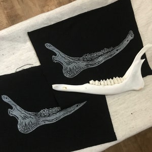 May include: Two black fabric patches with a white printed image of a jawbone with teeth. A white bone jawbone with teeth is lying on top of the fabric patches.