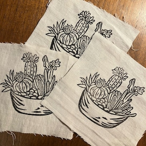 May include: Three square pieces of light-colored fabric with a black linocut print of a cactus garden in a basket. The cactus garden includes various cacti and succulents. The fabric has a raw, unfinished edge.
