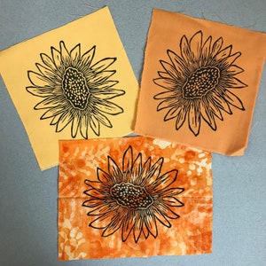 May include: Three fabric squares with black sunflower designs. The squares are in shades of yellow and orange.