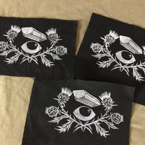 May include: Three black fabric patches with white embroidered designs. Each patch features a stylized eye with a cross, a crystal, and a cluster of thistle flowers.